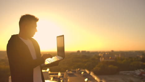 A-male-freelance-programmer-sits-on-a-skyscraper-roof-with-a-laptop-and-beer-typing-code-on-a-keyboard-during-sunset.-Remote-work